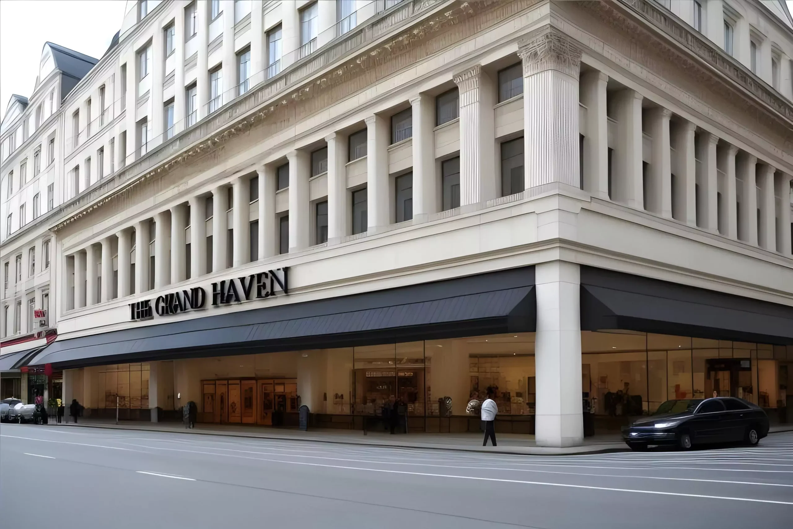 Exterior of a grand retail building named 'The Grand Haven,' featuring neoclassical architecture with tall columns and large glass windows, situated along a city street with a parked car and pedestrians.