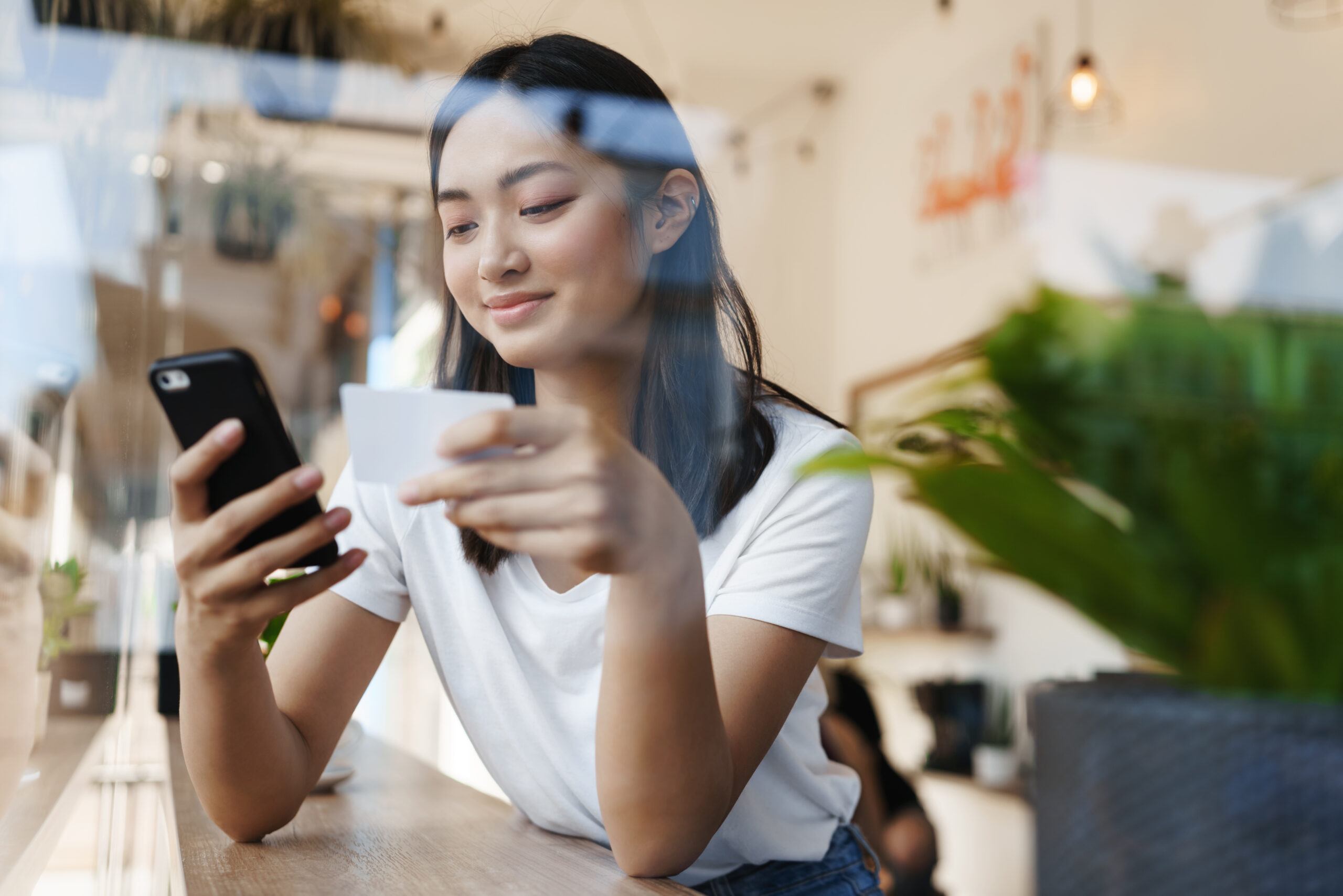 Stylish asian girl sitting in cafe near window, paying for online shopping with credit card, transfer money for coffee with smartphone, casually sitting in restaurant on summer day.